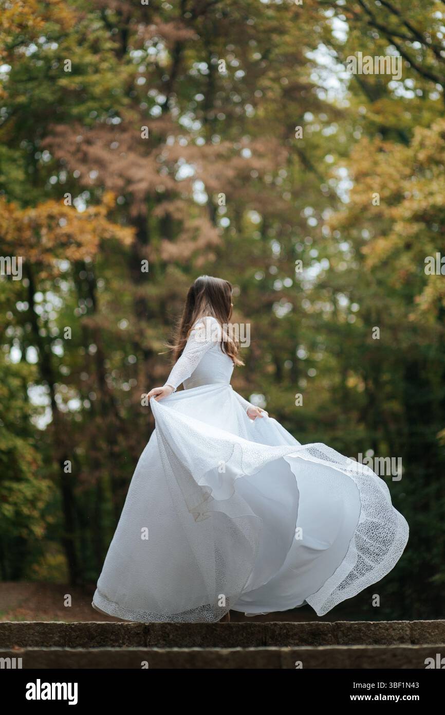 Una sposa si intreccia gioiosamente in una foresta, il suo abito fluente e il copricapo floreale aggiungono un'atmosfera eterea e incantevole. Foto Stock