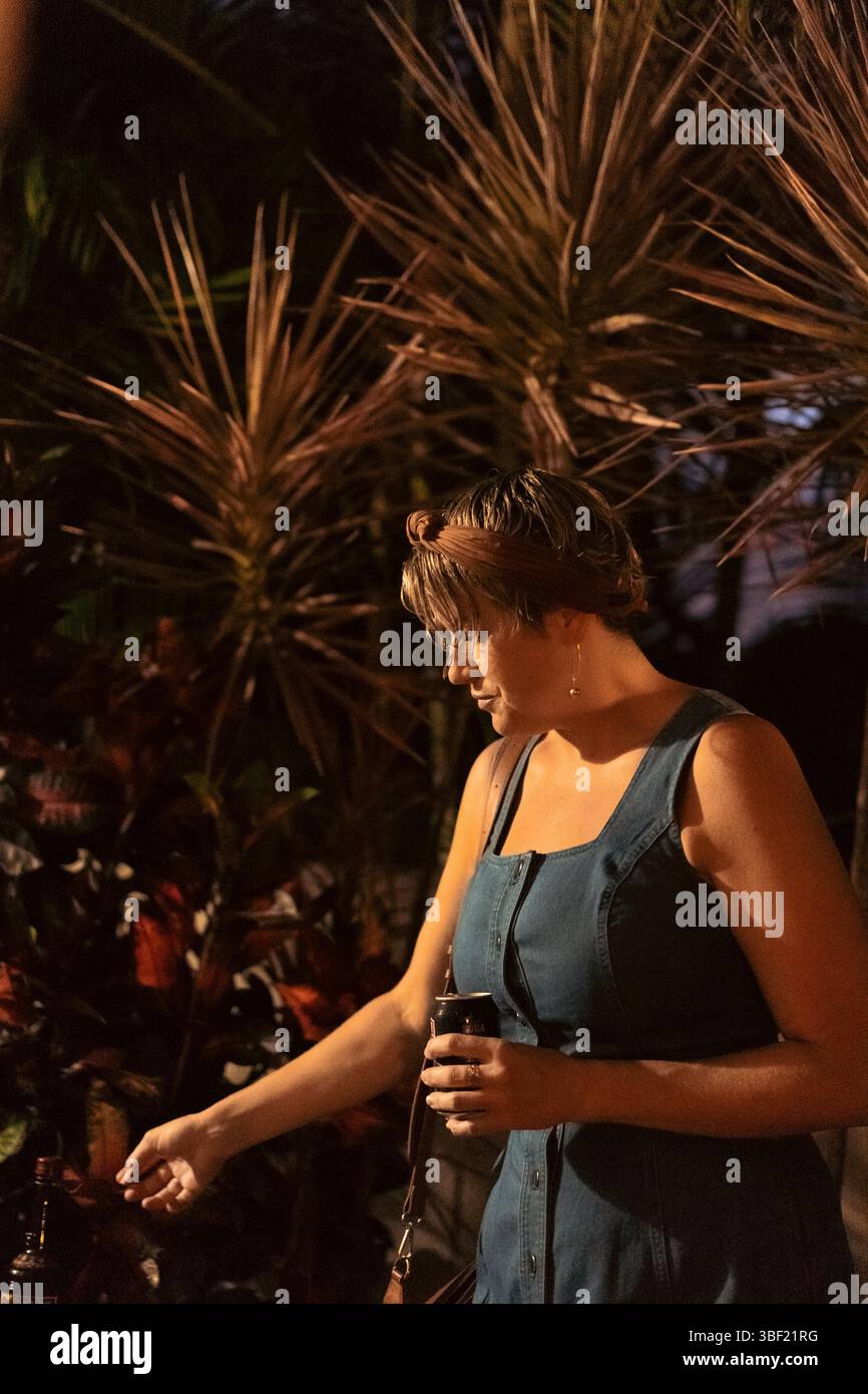 Donna in abito denim che tiene un drink durante una riunione notturna all'aperto Foto Stock