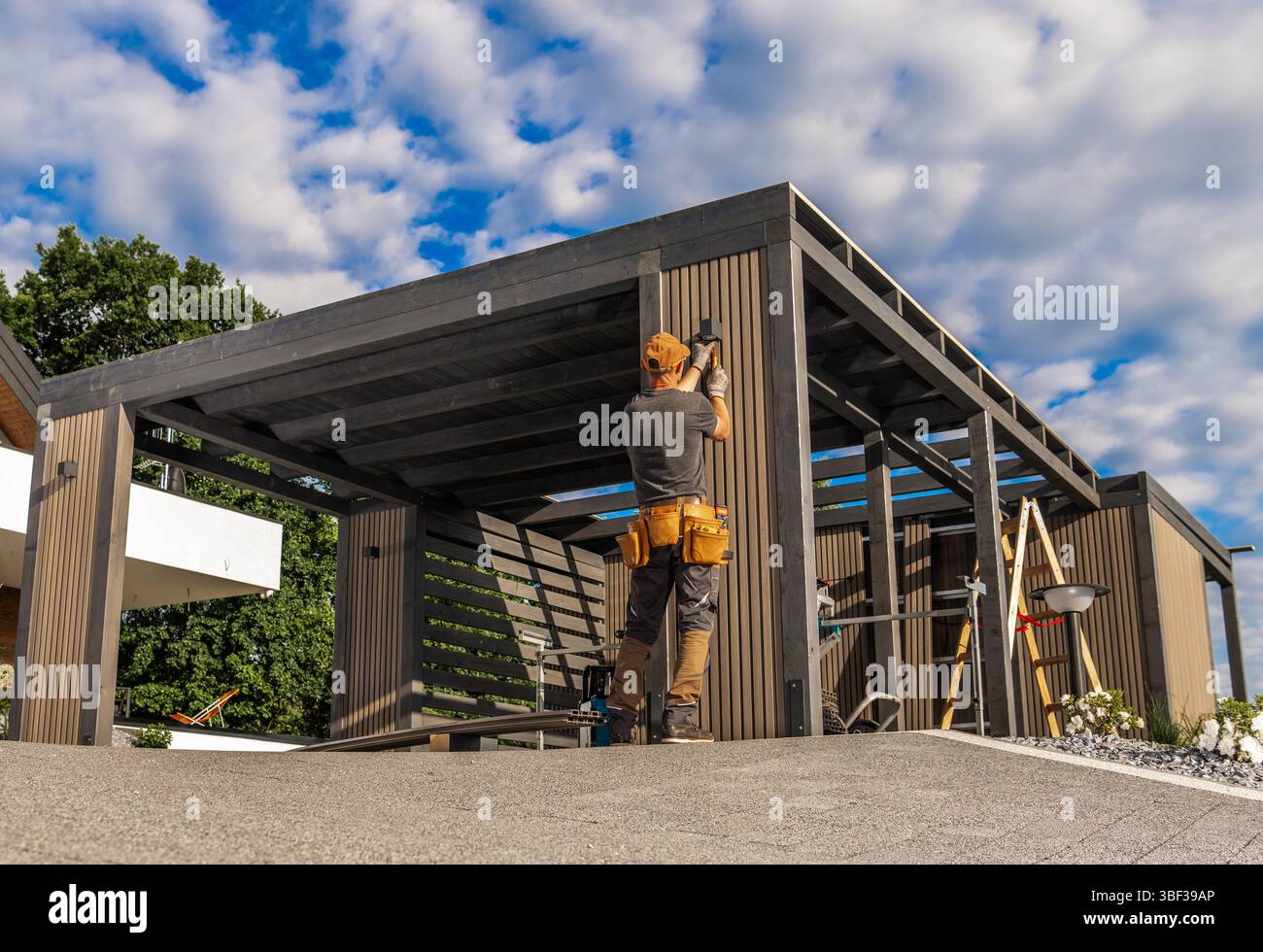 Un lavoratore esperto sta costruendo una struttura esterna, concentrandosi su dettagliate cornici in legno sotto un cielo luminoso con nuvole. L'area presenta un moderno resid Foto Stock