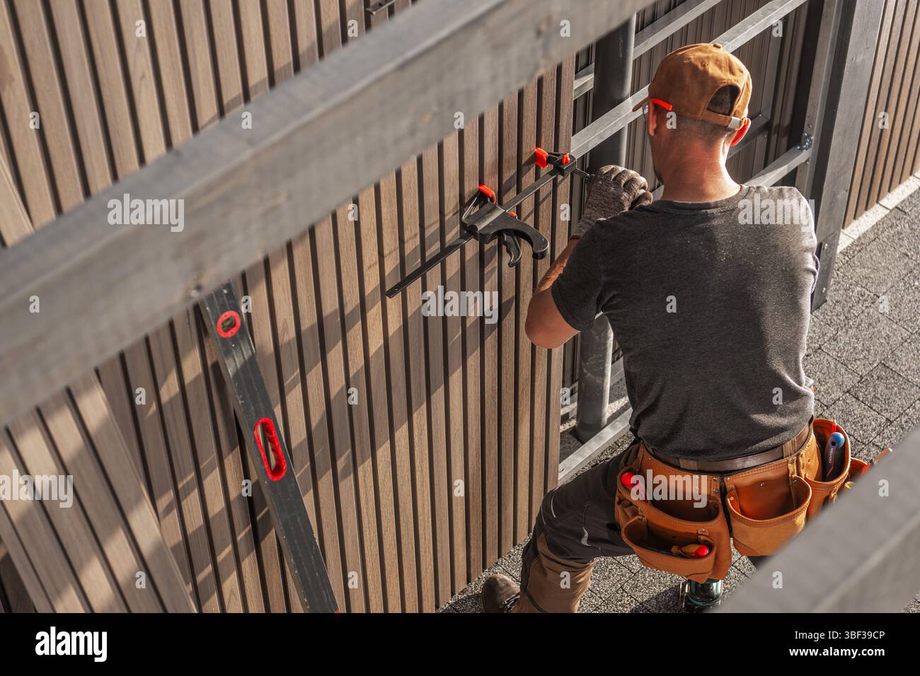 Un lavoratore esperto si concentra sull'installazione di pannelli in legno di lamella all'esterno di un edificio. Il sole proietta ombre, evidenziando gli strumenti e i materiali circostanti Foto Stock