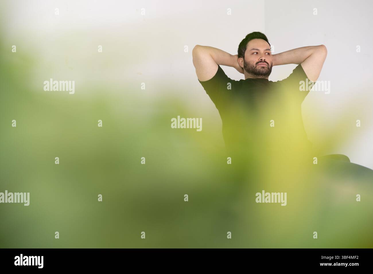 Un uomo calmo si appoggia con le mani dietro la testa, parzialmente nascosto dietro una morbida sfocatura delle piante, evocando pace, pause e momenti introspettivi. Foto Stock