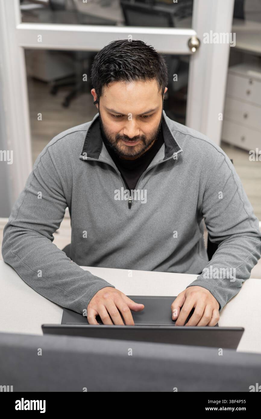 Un uomo con barba e blazer grigio sul suo laptop, rivolto in avanti con concentrazione. L'ambiente moderno dell'ufficio mette in risalto produttività e professionalità Foto Stock