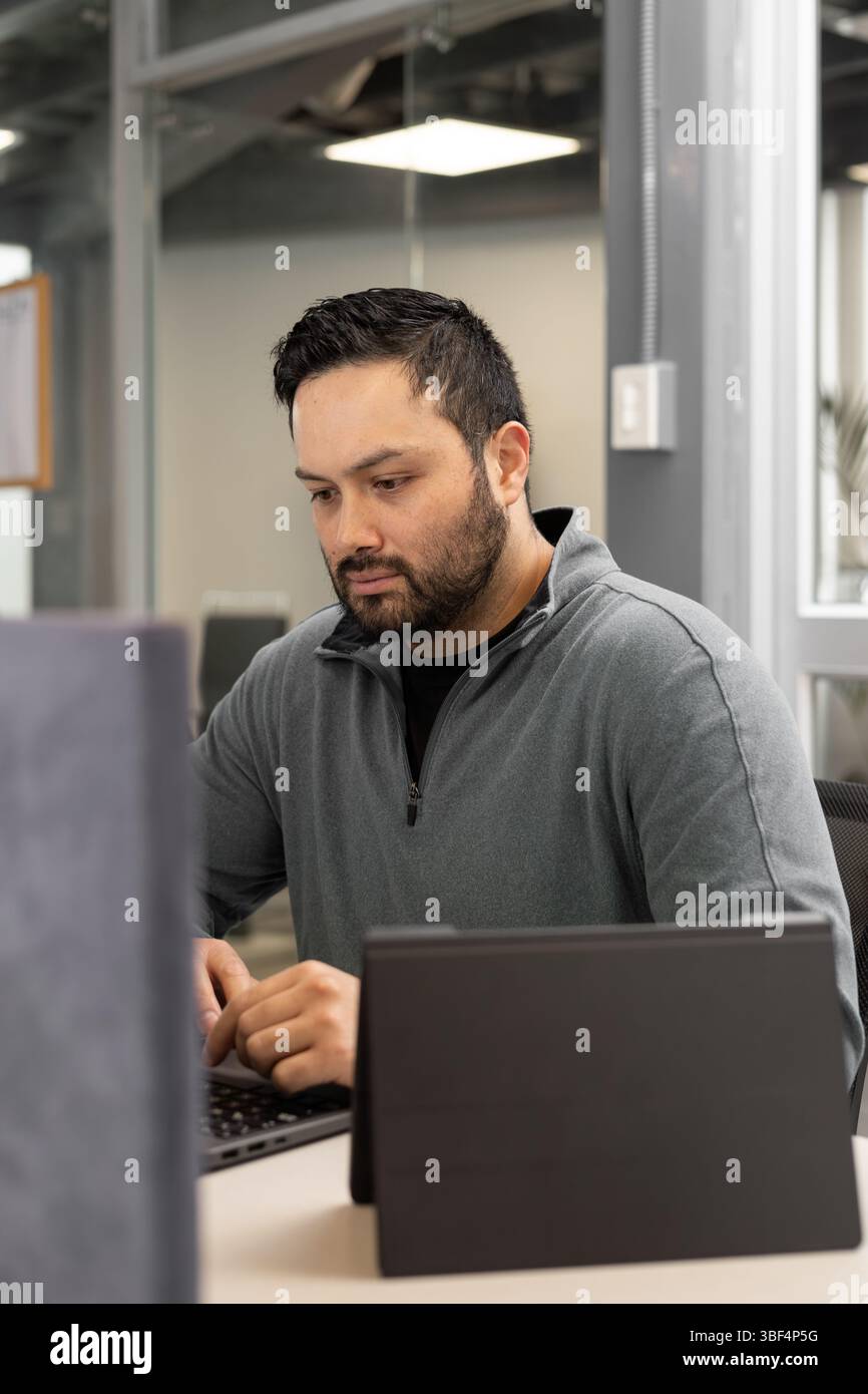Un uomo con la barba e il blazer grigio si siede alla sua scrivania, guardando il suo notebook con un'espressione premurosa. La scena dell'ufficio suggerisce riflessioni, pianificazioni Foto Stock