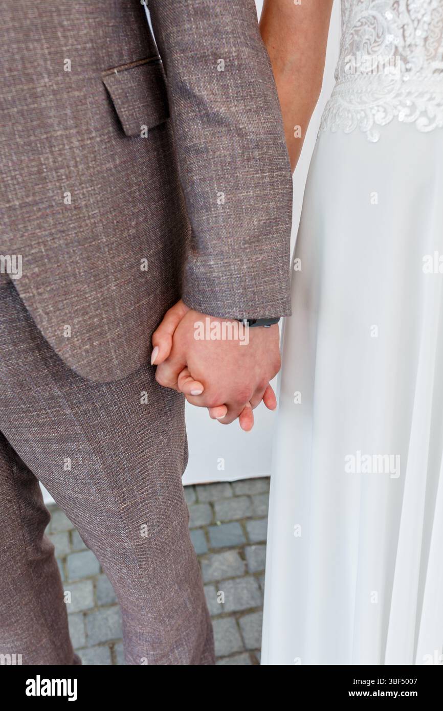 Uno scatto ravvicinato mostra una coppia di sposi che si tiene per mano durante la cerimonia di nozze, con lo sposo che indossa un abito marrone in tweed e la sposa in bianco Foto Stock
