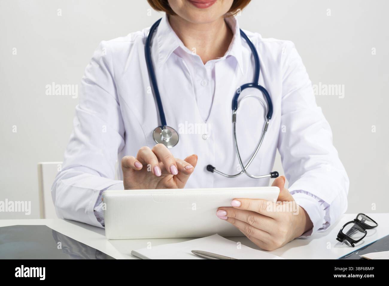 Avvicinare le mani di un medico con un tablet. Medico di famiglia in camice bianco con stetoscopio seduto alla scrivania. Diagnosi medica professionale AN Foto Stock