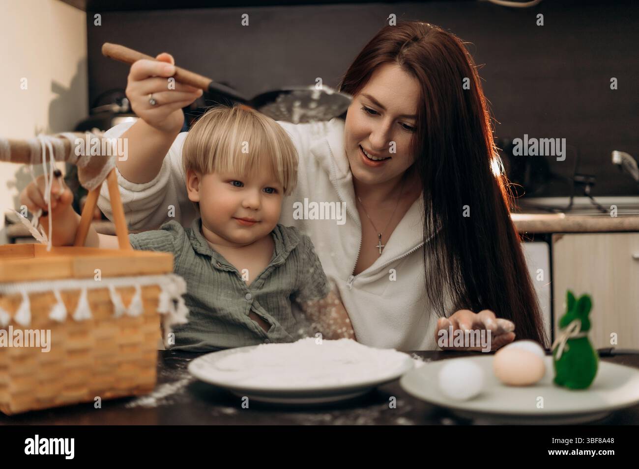 La genitorialità creativa incontra il divertimento pasquale al coperto, mentre il bambino esplora la farina con un gioco sensoriale, un apprendimento gioioso e le tradizioni di famiglia in un momento accogliente in cucina Foto Stock