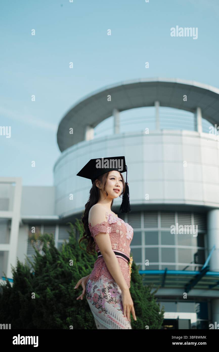 Orgogliosa donna del Sud-est asiatico laureata in abiti tradizionali di fronte al moderno edificio del campus Foto Stock