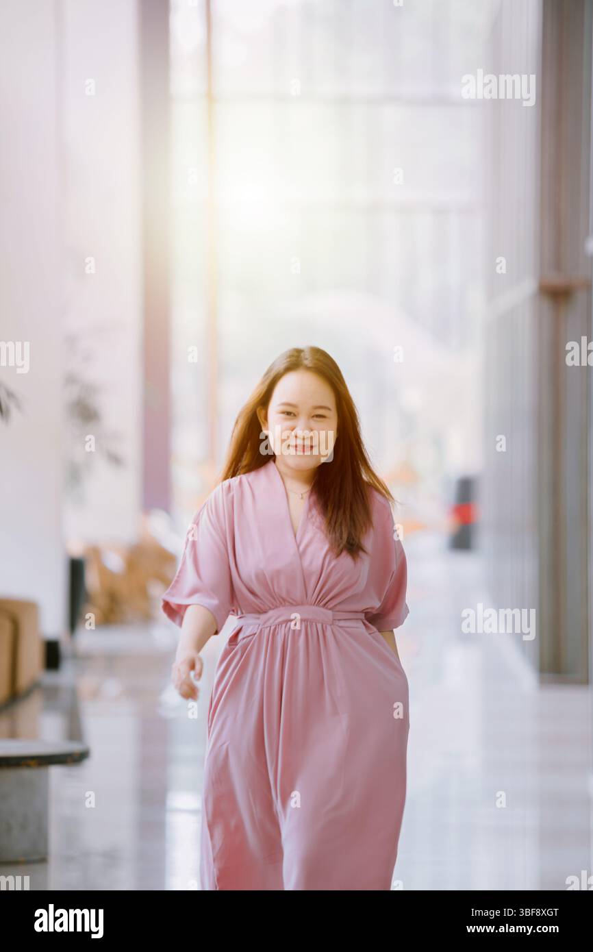Donna sorridente del sud-est asiatico in abito rosa che cammina tranquillamente in un moderno corridoio illuminato dal sole con espressione rilassata e sfondo bokeh Foto Stock