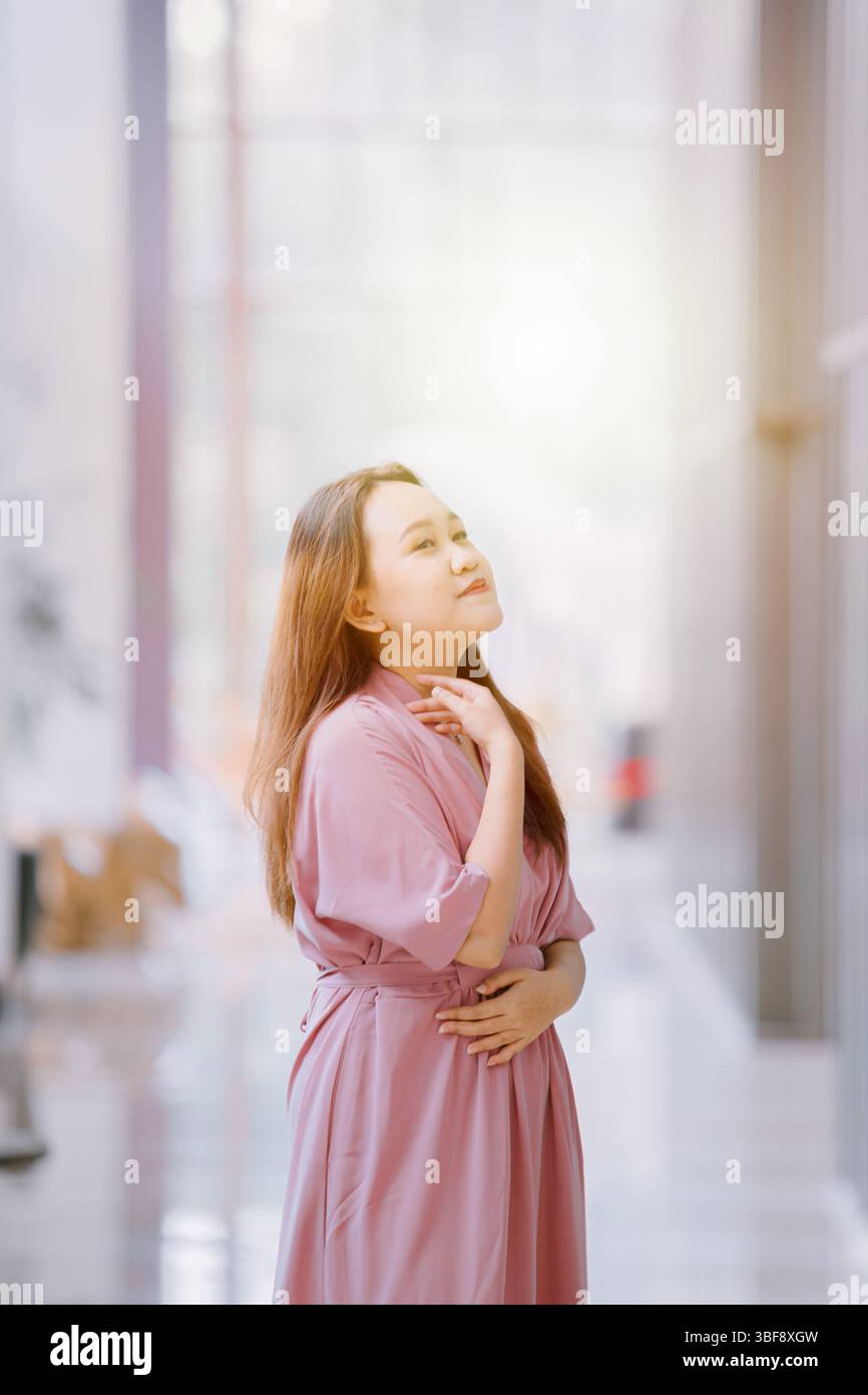 Elegante donna del sud-est asiatico in abito rosa in un corridoio luminoso con luce solare sullo sfondo bokeh Foto Stock