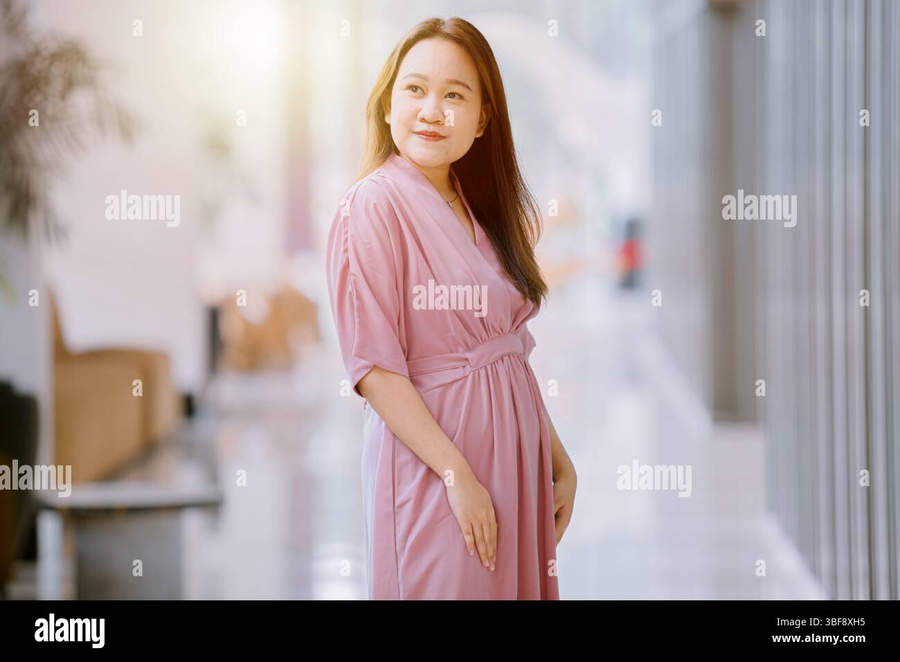 Elegante donna del sud-est asiatico in abito rosa in un corridoio luminoso con luce solare sullo sfondo bokeh Foto Stock