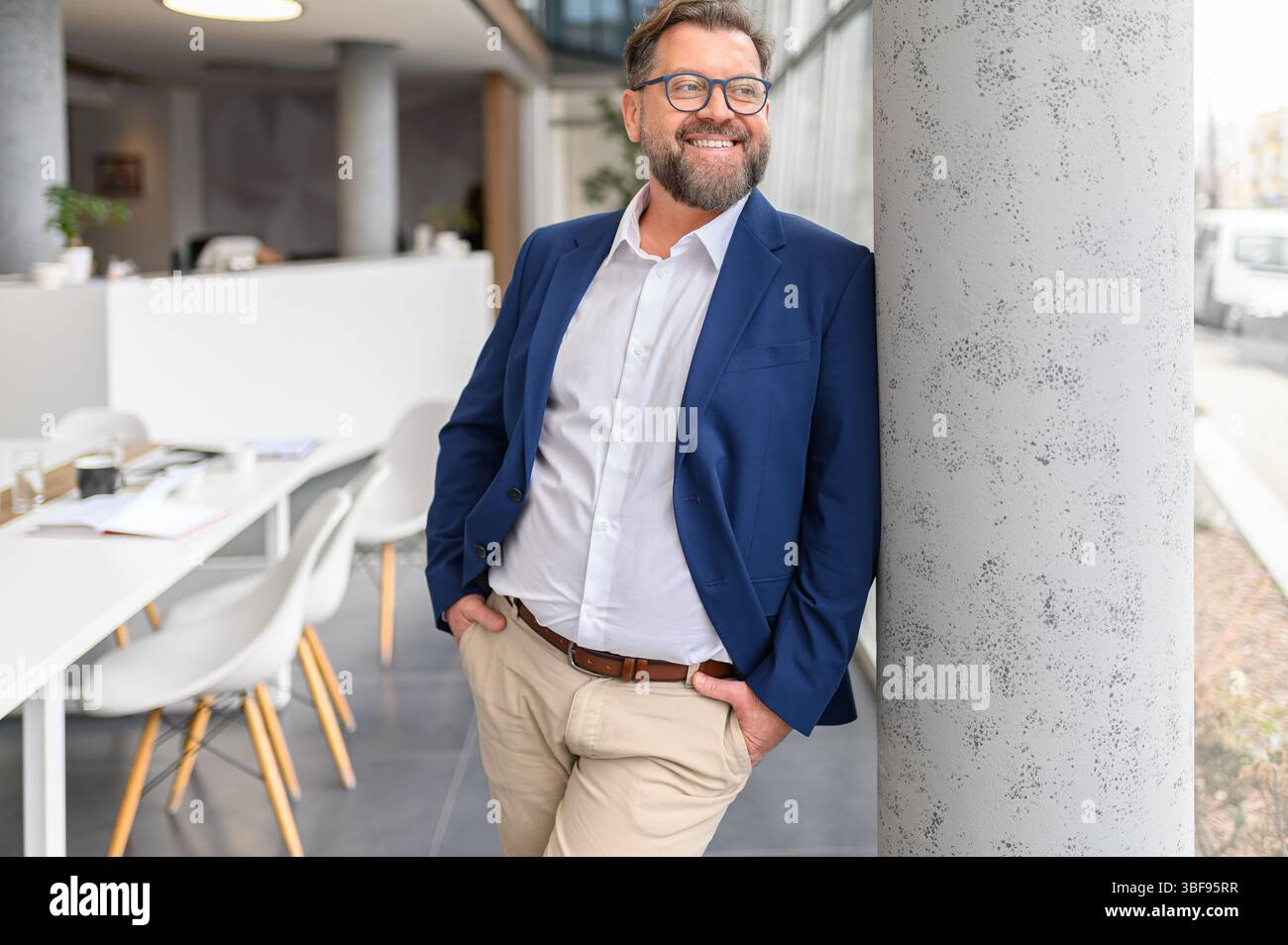 Ritratto di un manager sorridente in occhiali da vista e con le mani in tasca appoggiate sulla colonna in un ufficio moderno Foto Stock
