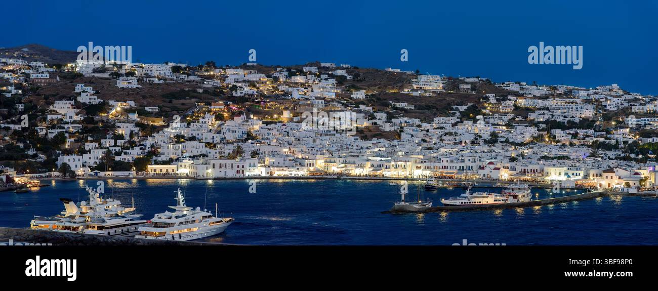 Il porto di Mykonos di notte, la Grecia, il vivace porto di Mykonos, brulicante di barche da pesca, yacht eleganti e vivaci caffetterie sul lungomare. Gli edifici imbiancati brillano sotto il sole dell'Egeo, mentre la brezza salata trasporta il ronzio della conversazione e il profumo del mare, una scena senza tempo dove tradizione ed energia cosmopolita si incontrano su questa iconica isola greca. Mykonos è un'isola del gruppo delle Cicladi nel Mar Egeo. Foto Stock