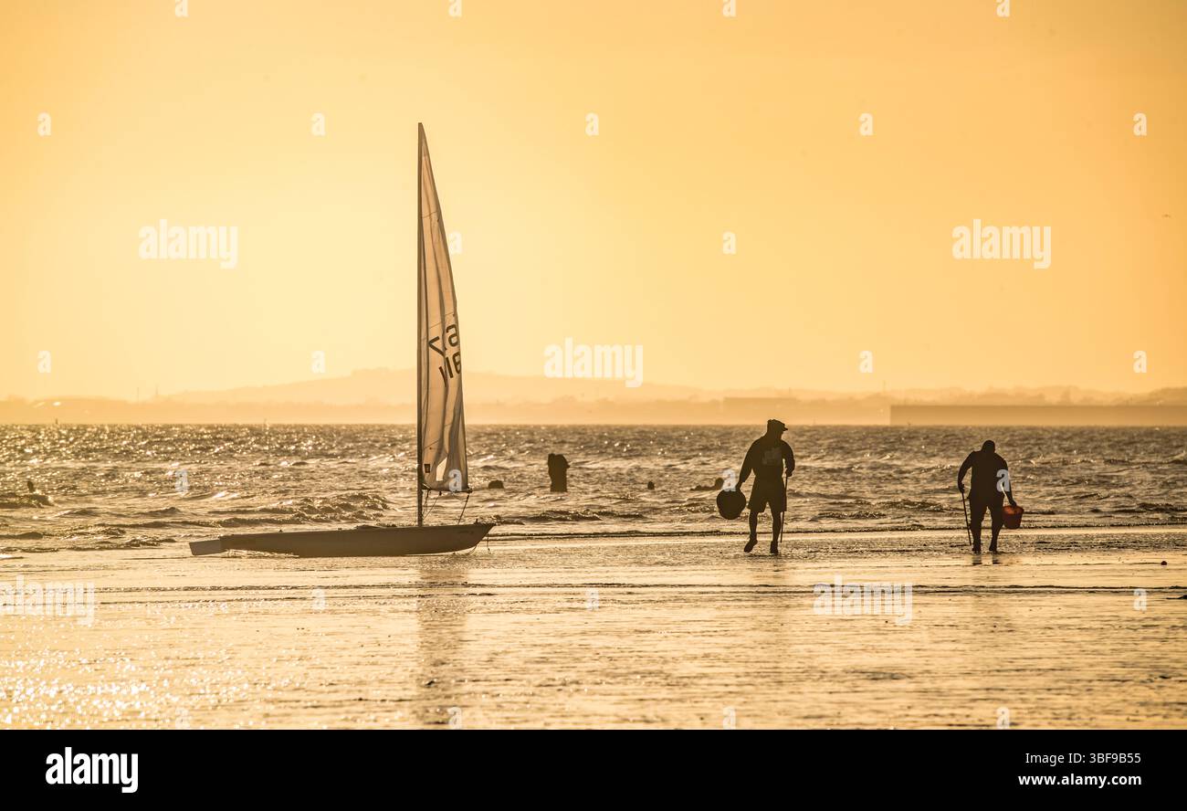 Pescatori con secchi che scavano nella sabbia con la bassa marea per i vermi da usare come esca. Una piccola barca a vela nel surf. Spiaggia di Brighton. Inghilterra. Silhouette. Brighton, Inghilterra, Regno Unito. Foto Stock