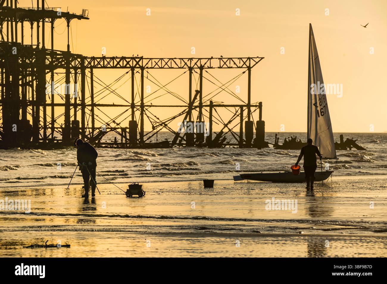 Pescatori con secchi che scavano nella sabbia con la bassa marea per i vermi da usare come esca. Una piccola barca a vela e la struttura abbandonata del West Pier sullo sfondo. Spiaggia di Brighton. Inghilterra. Silhouette. Brighton, Inghilterra, Regno Unito. Foto Stock