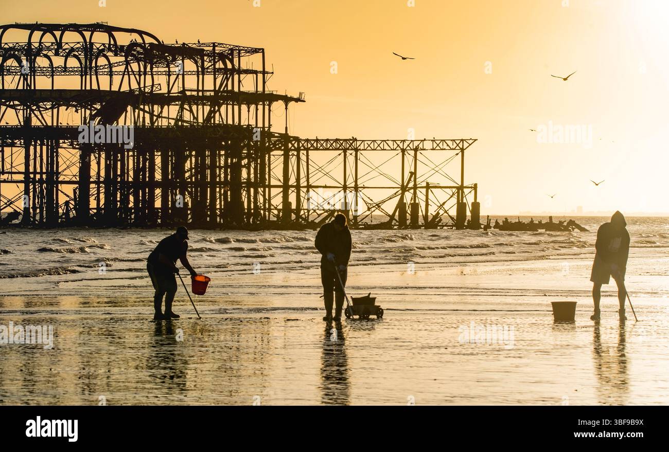 Pescatori con secchi che scavano nella sabbia con la bassa marea per i vermi da usare come esca. Una piccola barca a vela e la struttura abbandonata del West Pier sullo sfondo. Spiaggia di Brighton. Inghilterra. Silhouette. Brighton, Inghilterra, Regno Unito. Foto Stock