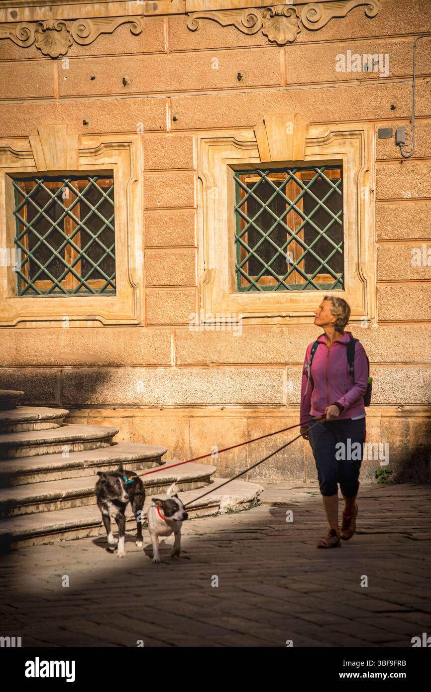 Donna che cammina due cani in porta in via Roma, oltre alla piazza con gradini della chiesa di San Giovanni Battista alla sua destra, finale Ligure, Italia. Foto Stock