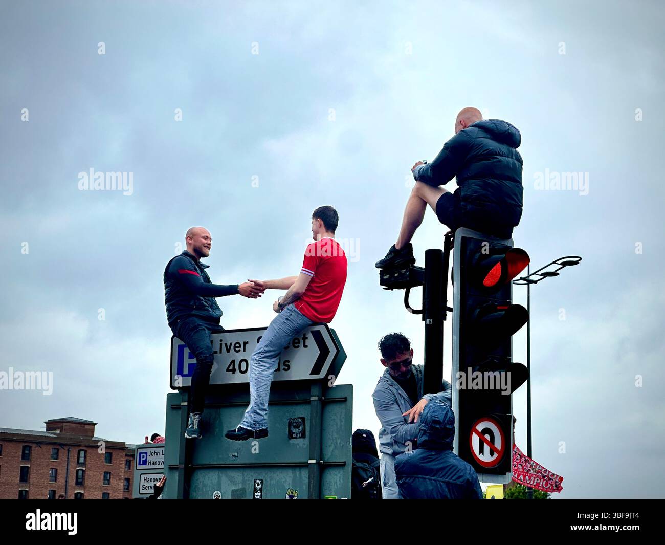 Gli uomini stringono le mani sulla cima dei cartelli stradali mentre i fan scalano i semafori sullo Strand durante la parata della vittoria di Liverpool, il 25 maggio 2025 - Immagine stock catturata con smartphone