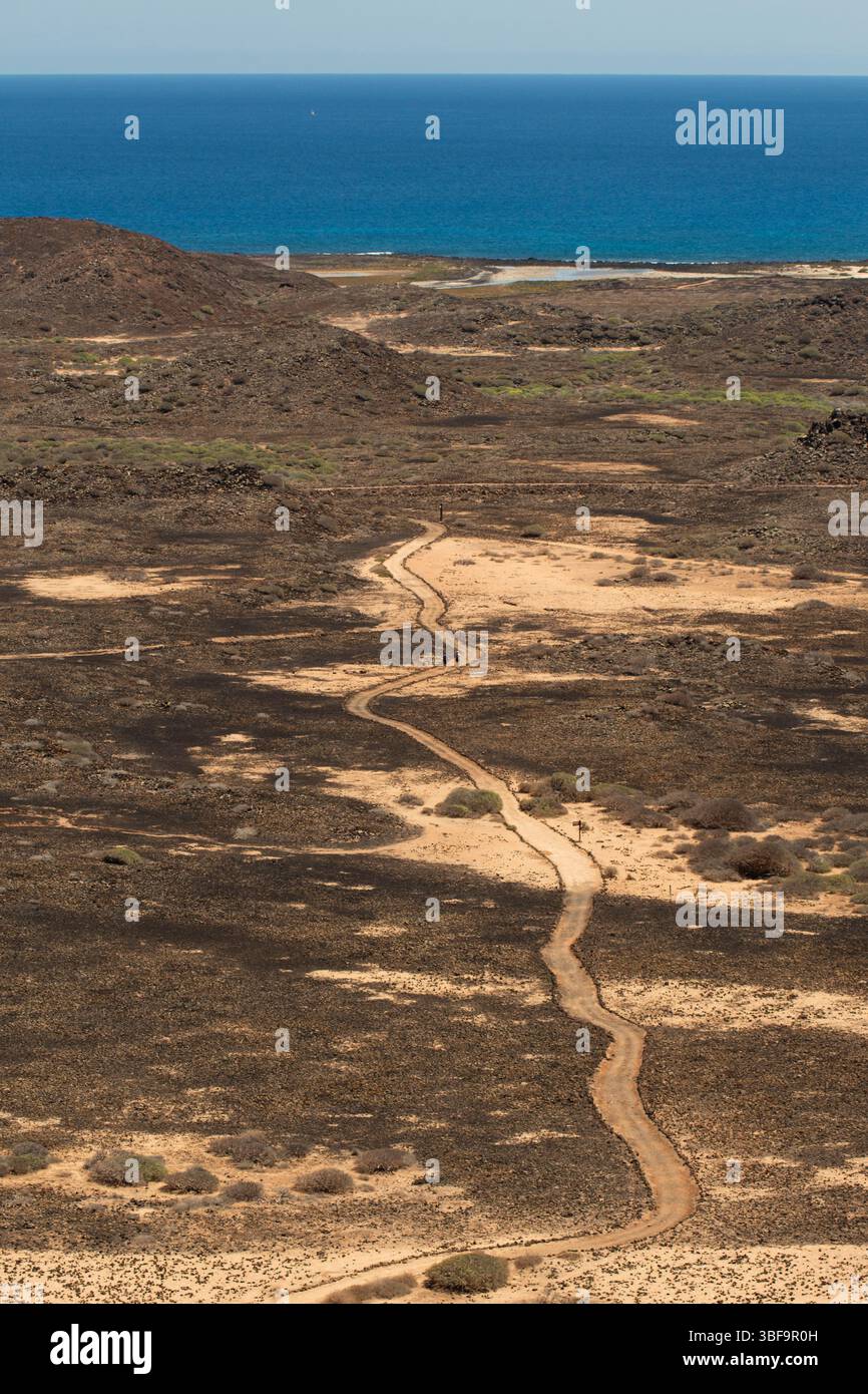 Foto in stile aereo di escursionisti che camminano su un sentiero curvilineo attraverso il terreno vulcanico verso il mare. La scena evidenzia scala, vastità, isolamento e prog Foto Stock