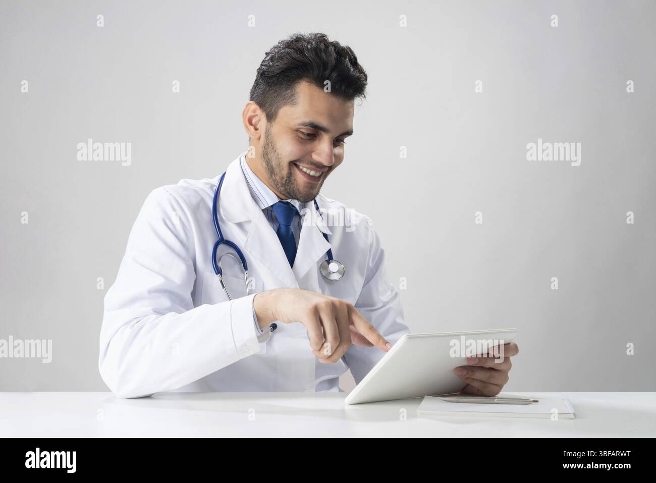 Giovane internista sorridente che usa un tablet. Serio terapista in abito medico bianco con stetoscopio seduto alla scrivania. Tecnologia digitale moderna in Foto Stock