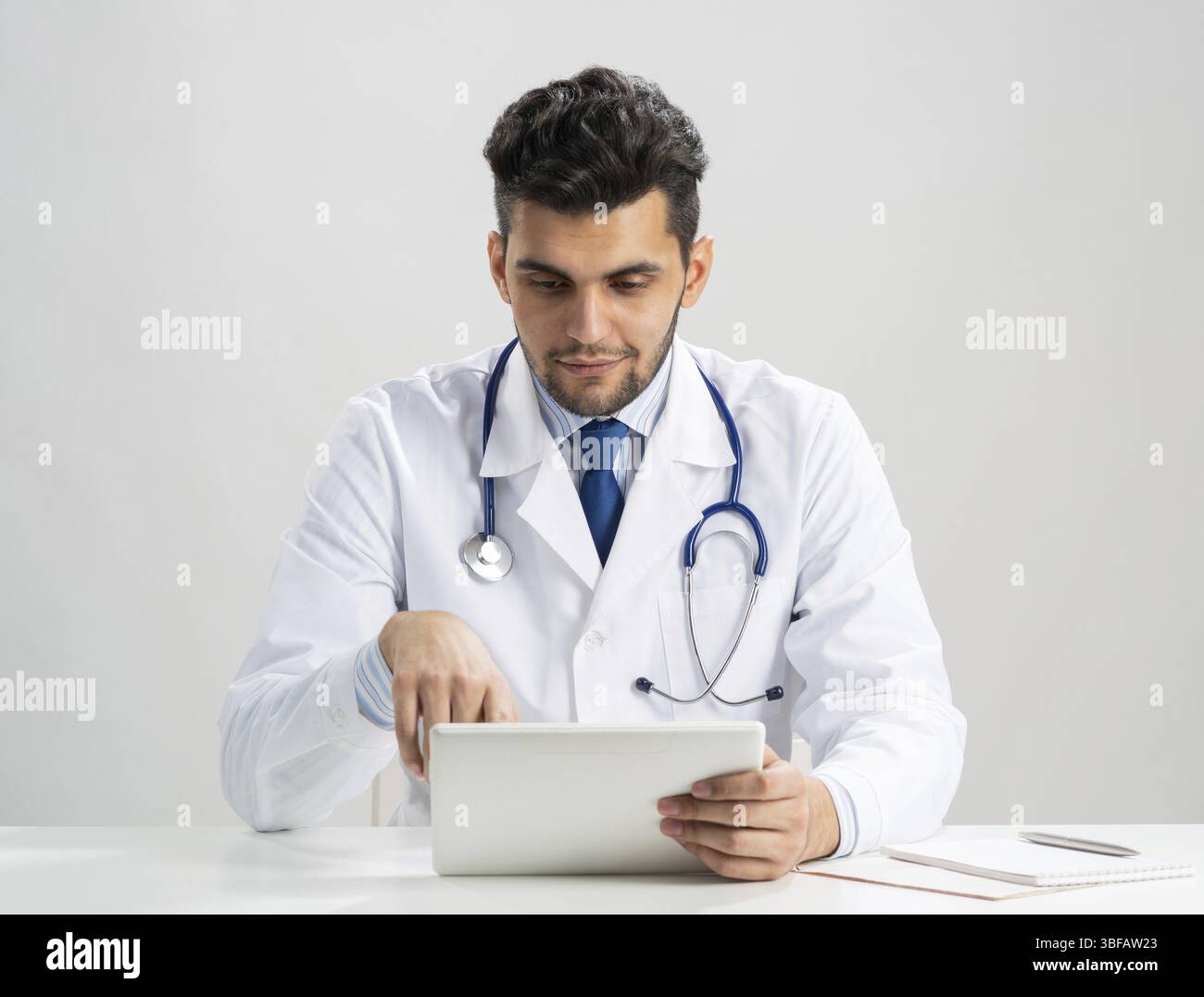 Giovane internista arabo che usa un tablet. Serio terapista in abito medico bianco con stetoscopio seduto alla scrivania. Moderna tecnologia digitale in diam Foto Stock