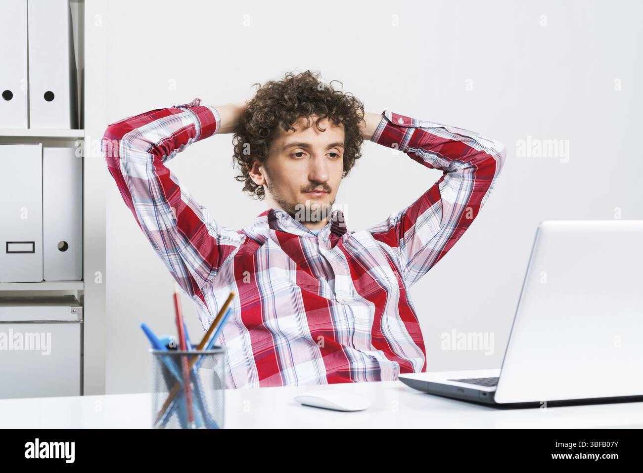 Giovane uomo rilassato in camicia controllata nel suo ufficio con mani sulla testa Foto Stock