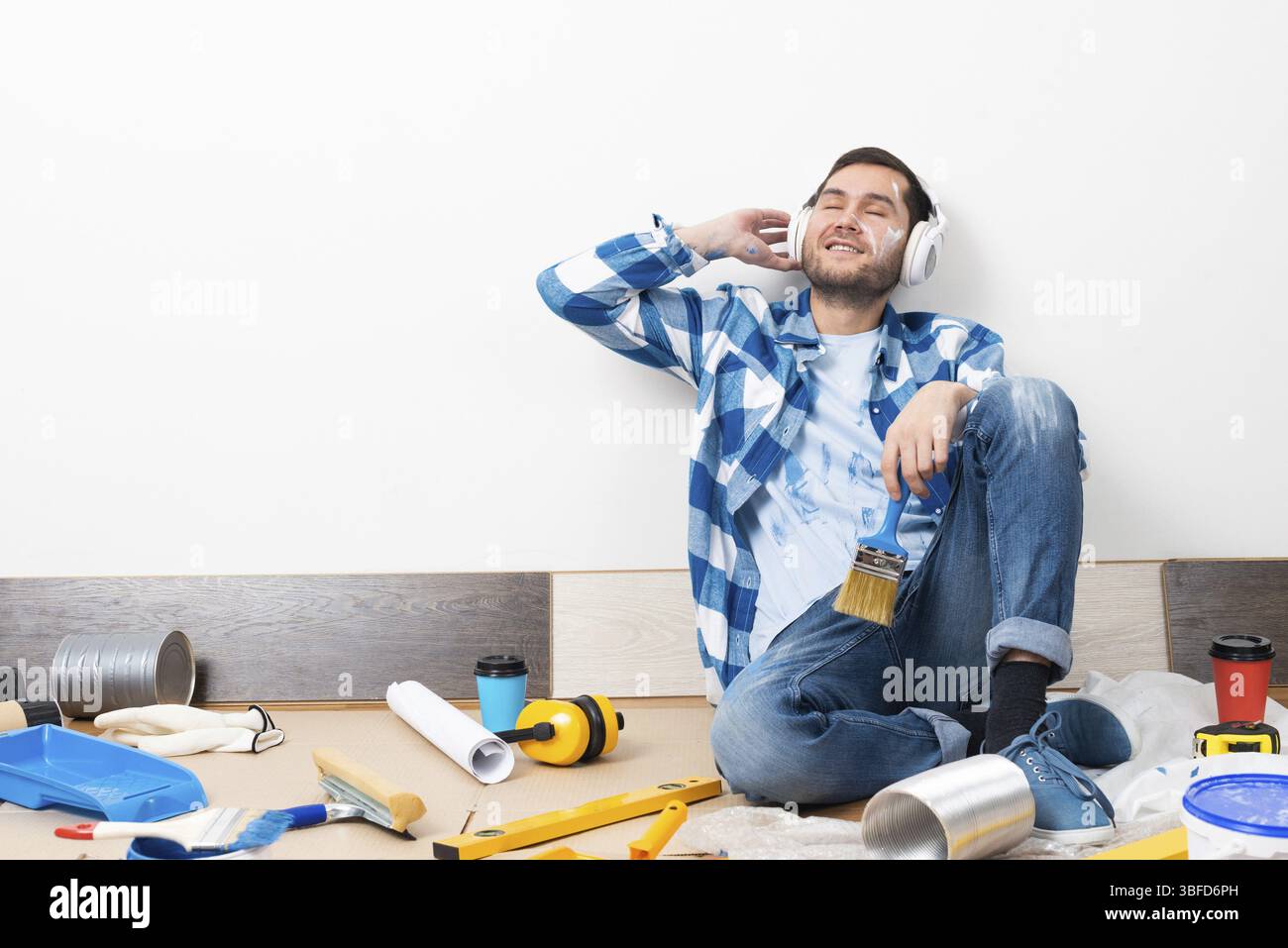 Bell'uomo barbuto che ascolta la musica con le cuffie a casa. Rimodellamento della casa dopo il concetto di trasloco. Strumenti di verniciatura e materiale sul pavimento. Giovane cauca Foto Stock