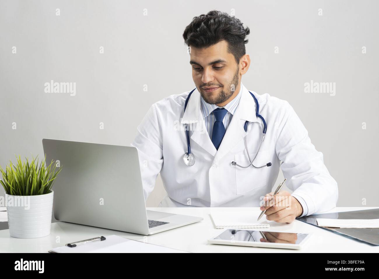 Giovane internista arabo che lavora al computer. Medico sorridente in camice bianco con stetoscopio seduto alla scrivania con computer portatile. Consulenza medica professionale Foto Stock