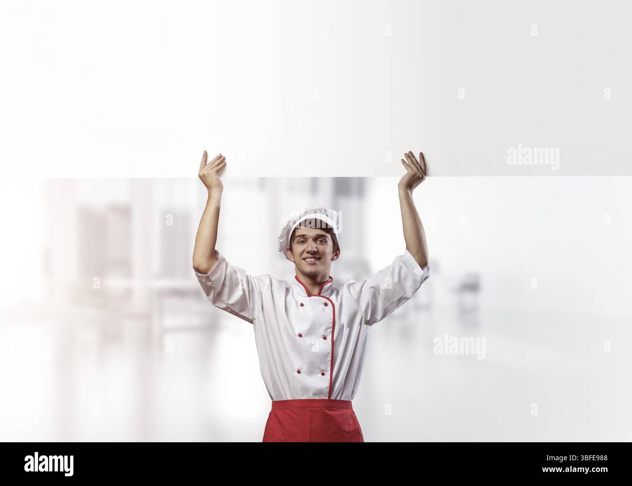 Giovane chef maschile con un cartello vuoto sopra la testa. Chef in uniforme bianca e rossa su sfondo sfocato in camera chiaro. Annuncio delle lezioni di cucina. Confessare Foto Stock
