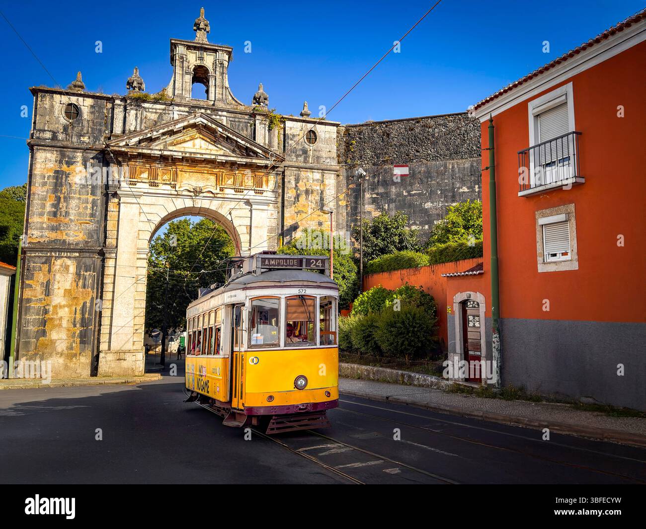 Lo storico tram giallo 24E di Lisbona passa attraverso un'arcata dell'antico acquedotto con colorati edifici portoghesi sotto il cielo azzurro del Portogallo Foto Stock