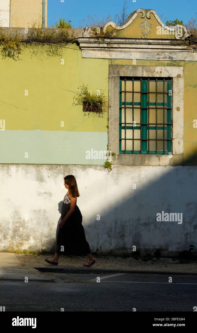 Una donna cammina davanti a pareti bianche e gialle intemprate con finestre verdi persiane che proiettano spettacolari ombre sulla strada acciottolata nella vecchia Lisbona Foto Stock