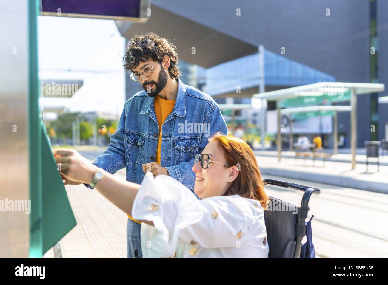 Donna in sedia a rotelle che convalida il suo biglietto presso una moderna fermata del tram, con un amico che fornisce assistenza e supporto durante il processo Foto Stock