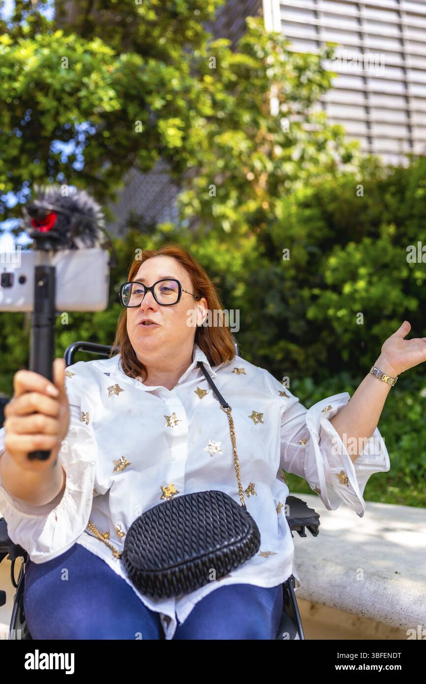 Creatore di contenuti Redhead seduto in sedia a rotelle per registrare video blog all'aperto utilizzando uno smartphone con microfono esterno Foto Stock