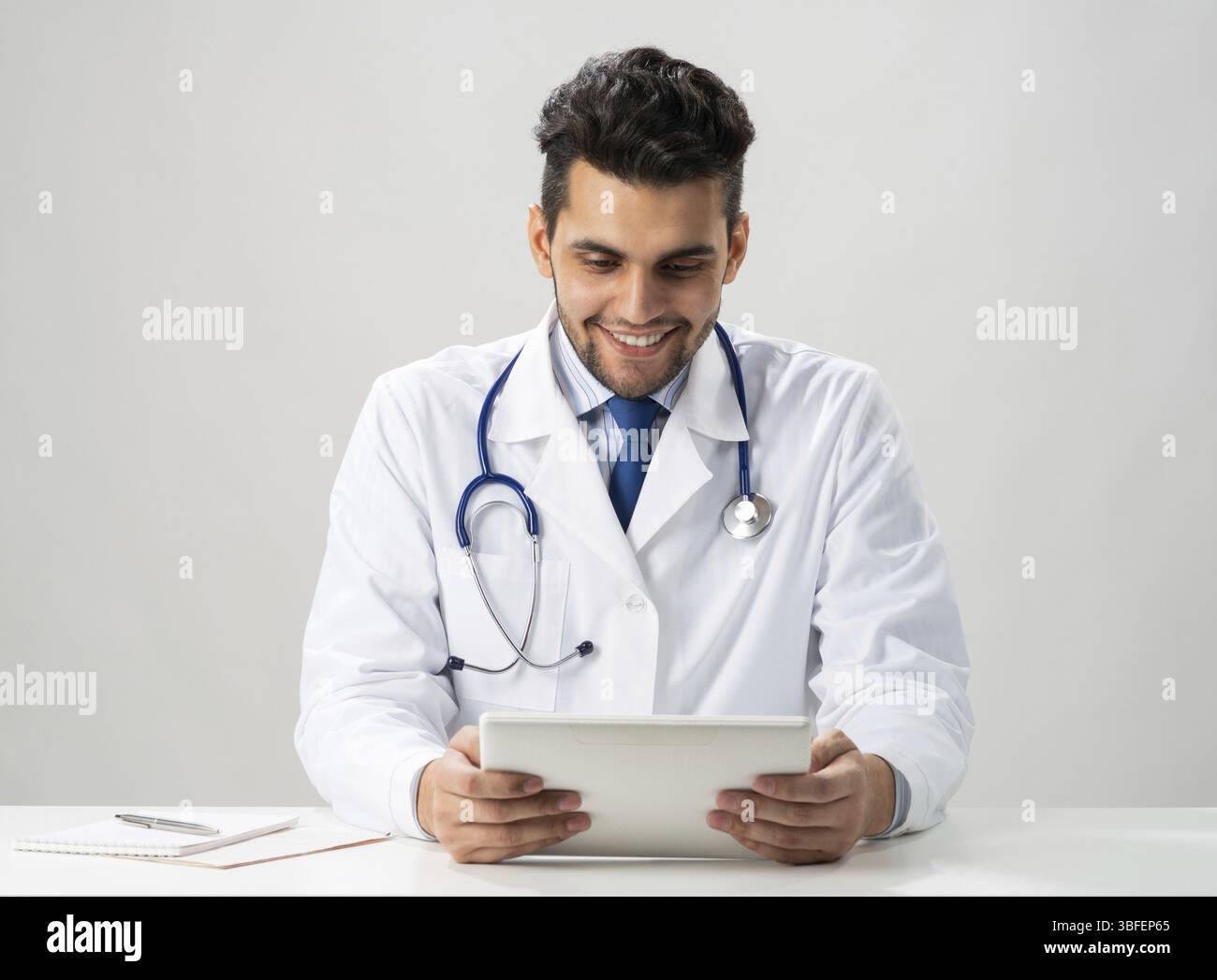 Giovane internista sorridente che usa un tablet. Serio terapista in abito medico bianco con stetoscopio seduto alla scrivania. Tecnologia digitale moderna in Foto Stock