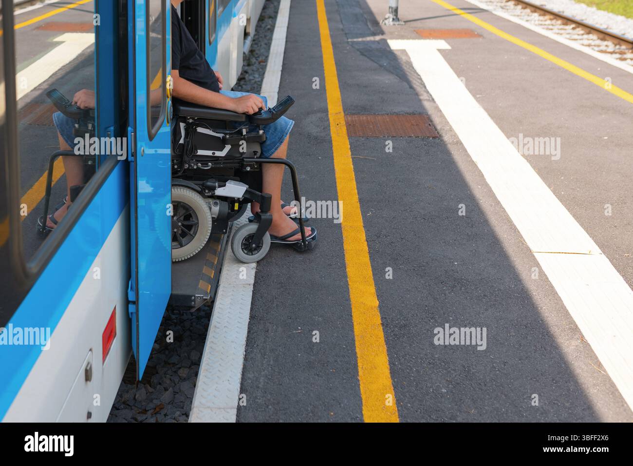 L'utente in sedia a rotelle che utilizza il treno accessibile attende su una piattaforma accanto a un treno. L'ambiente presenta linee chiare e una pista ben mantenuta Foto Stock