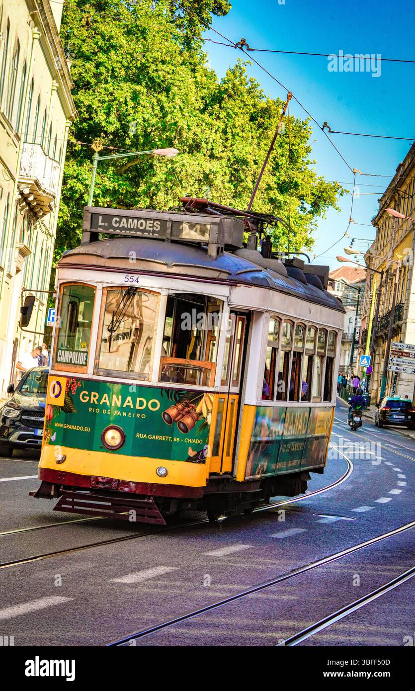 Il tram d'epoca 24E di Lisbona attraversa la strada alberata della città con cavi elettrici sospesi sotto il cielo azzurro soleggiato in Portogallo Foto Stock
