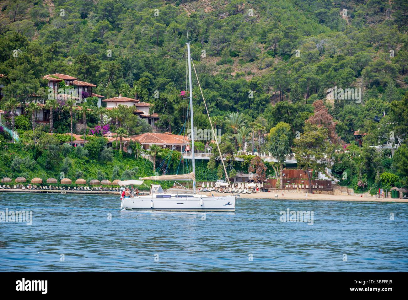 Un moderno yacht galleggiante vicino a una spiaggia privata, sostenuto da alberi vibranti e lussuose case vacanza, caratteristiche della costa turca. Foto Stock