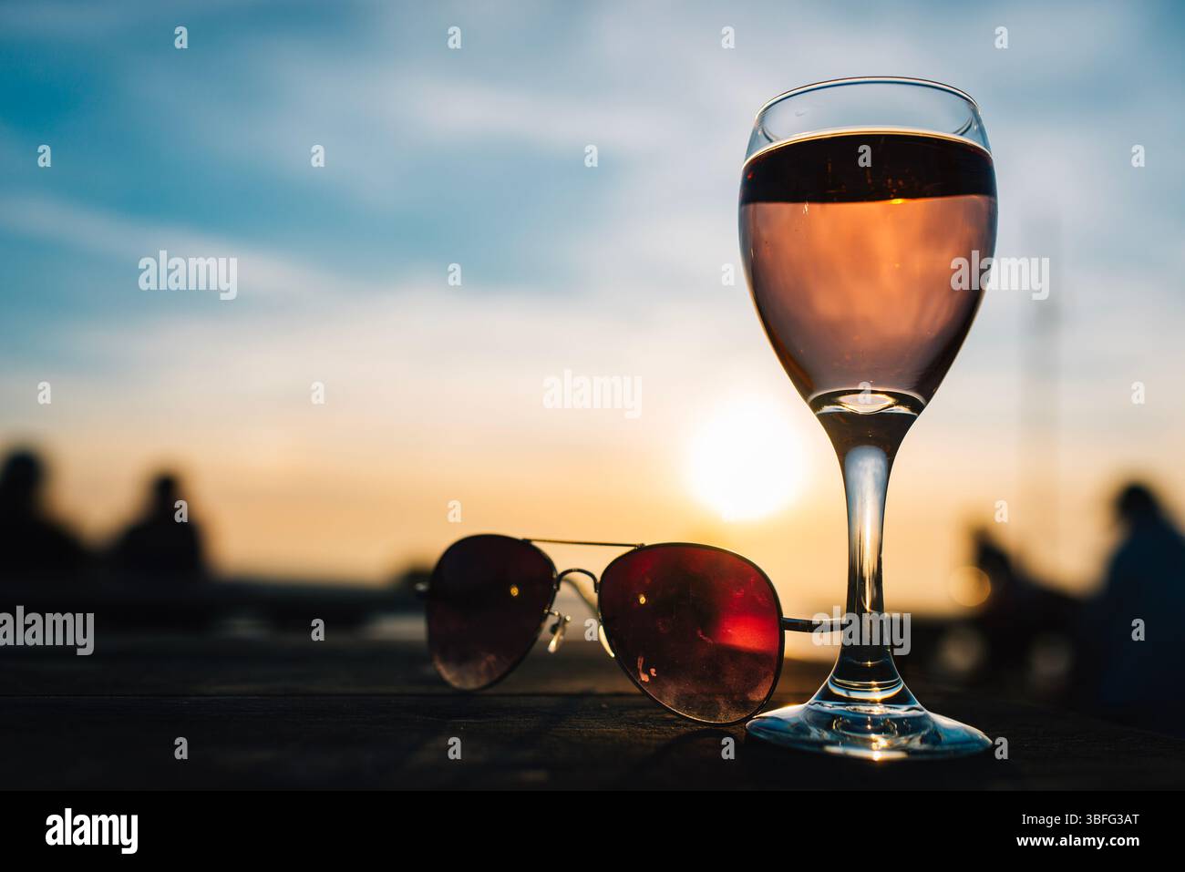 Bicchiere di rosé e occhiali da sole su un tavolo da bar sulla spiaggia al tramonto, catturando un rilassato umore estivo serale con tonalità calde e uno sfondo morbido. Foto Stock