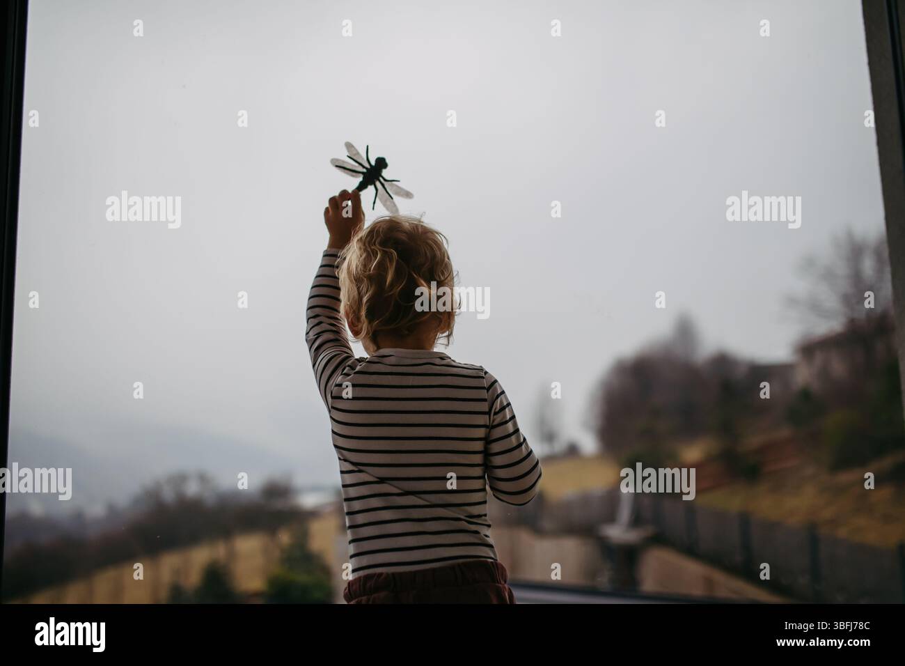 Un bambino adorabile sta giocando con il giocattolo delle libellule, in piedi accanto alla finestra. Foto Stock
