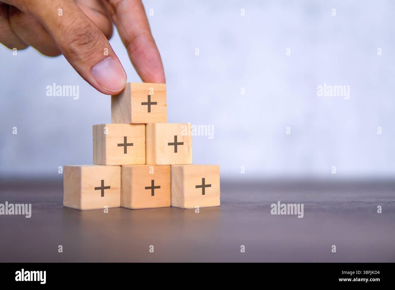 Una mano sta posizionando un cubo di legno con un segno più nero sulla cima di una piramide di cubi. Crescita, positiva, concetto di valore aggiunto. Foto Stock
