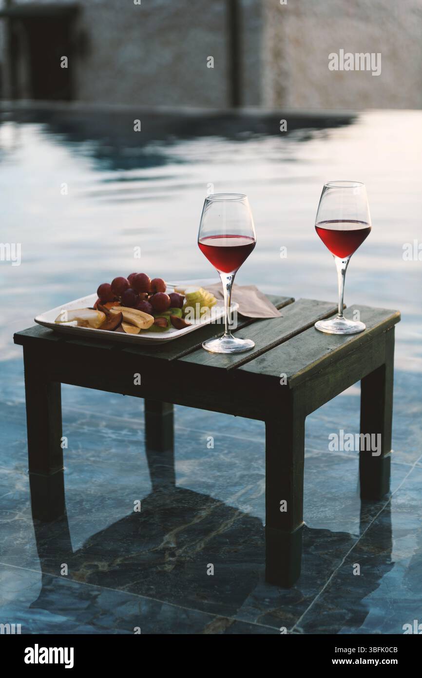Un intimo primo piano di due bicchieri da vino rosso e un vassoio di frutta organizzato ad arte a bordo piscina. Foto Stock