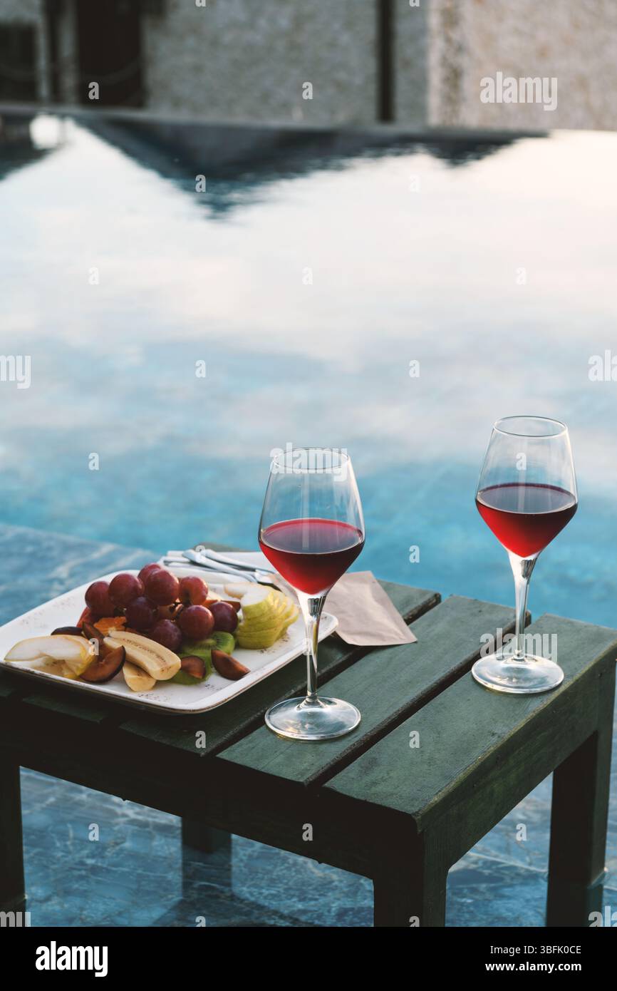 Un intimo primo piano di due bicchieri da vino rosso e un vassoio di frutta organizzato ad arte a bordo piscina. Foto Stock