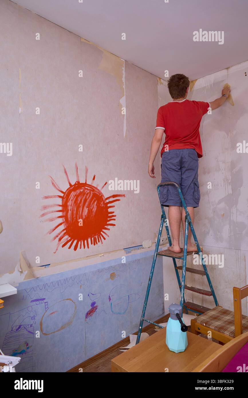Uomo in piedi su una scala che rimuove la vecchia carta da parati da una stanza. Disegni per bambini sbiaditi visibili e vernice rossa sul muro. La scena cattura la ristrutturazione della casa Foto Stock
