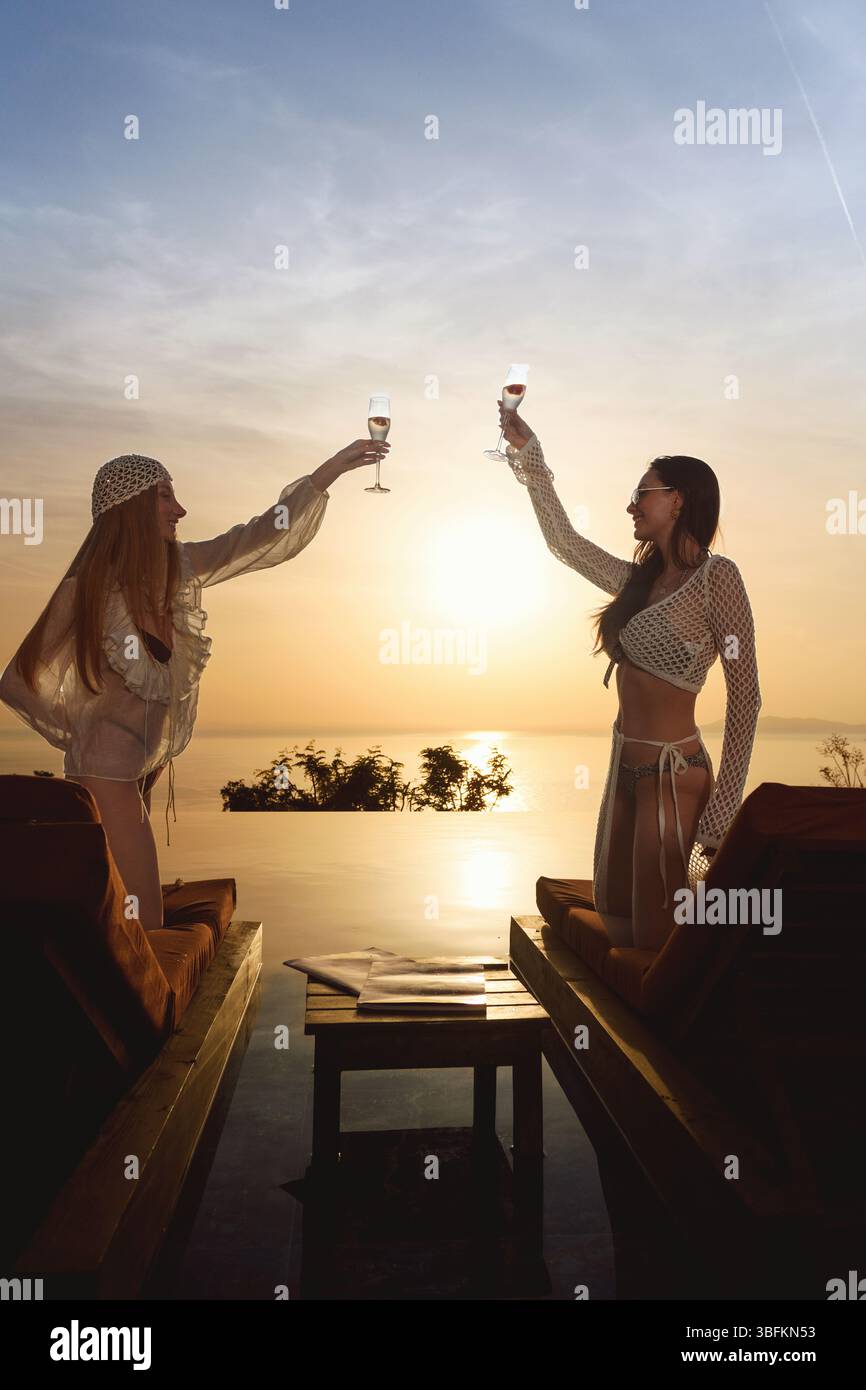 Una bruna e una bionda sui loro 20 bicchieri di champagne sui lettini da sole durante un pittoresco tramonto a bordo piscina. Foto Stock