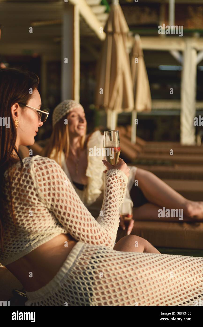 Una bruna e bionda nella loro lounge degli anni '20 sui lettini della piscina dell'hotel, gustando champagne durante l'ora dorata del tramonto. Foto Stock
