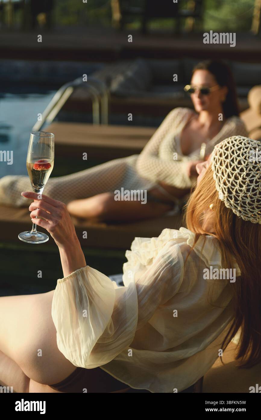 Una donna bruna degli anni '20 si rilassa su un lettino sorseggiando champagne durante un tramonto mozzafiato a bordo piscina. Foto Stock