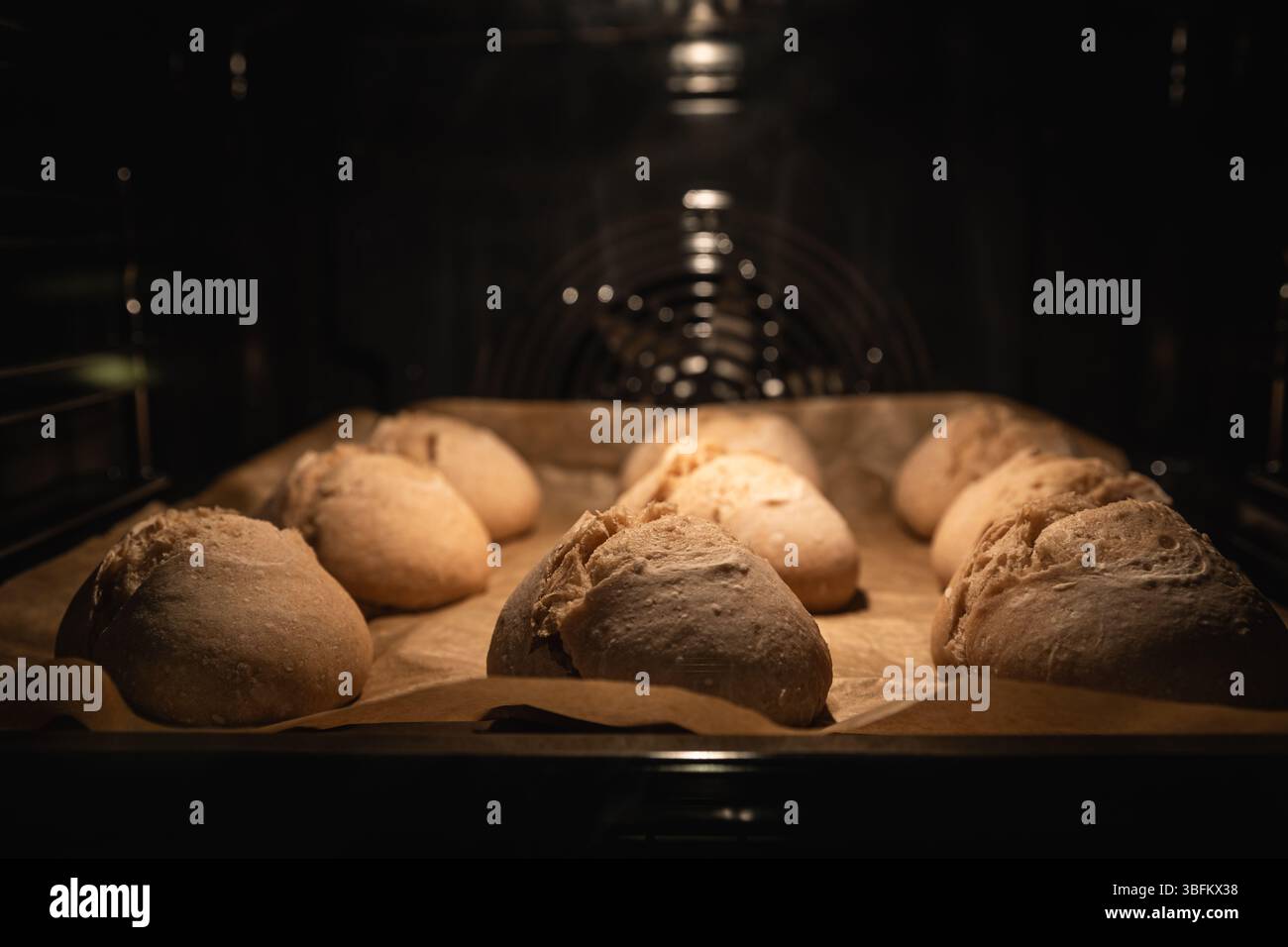 Primo piano di rustici rotoli di pasta madre su carta da forno, splendidamente innalzati con croste dorate. Calda atmosfera da cucina durante la cottura. Foto Stock