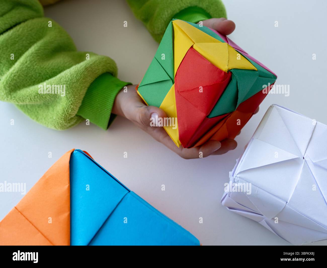 Bambino con maniche verdi che regge un luminoso cubo origami modulare sul tavolo bianco. Foto Stock