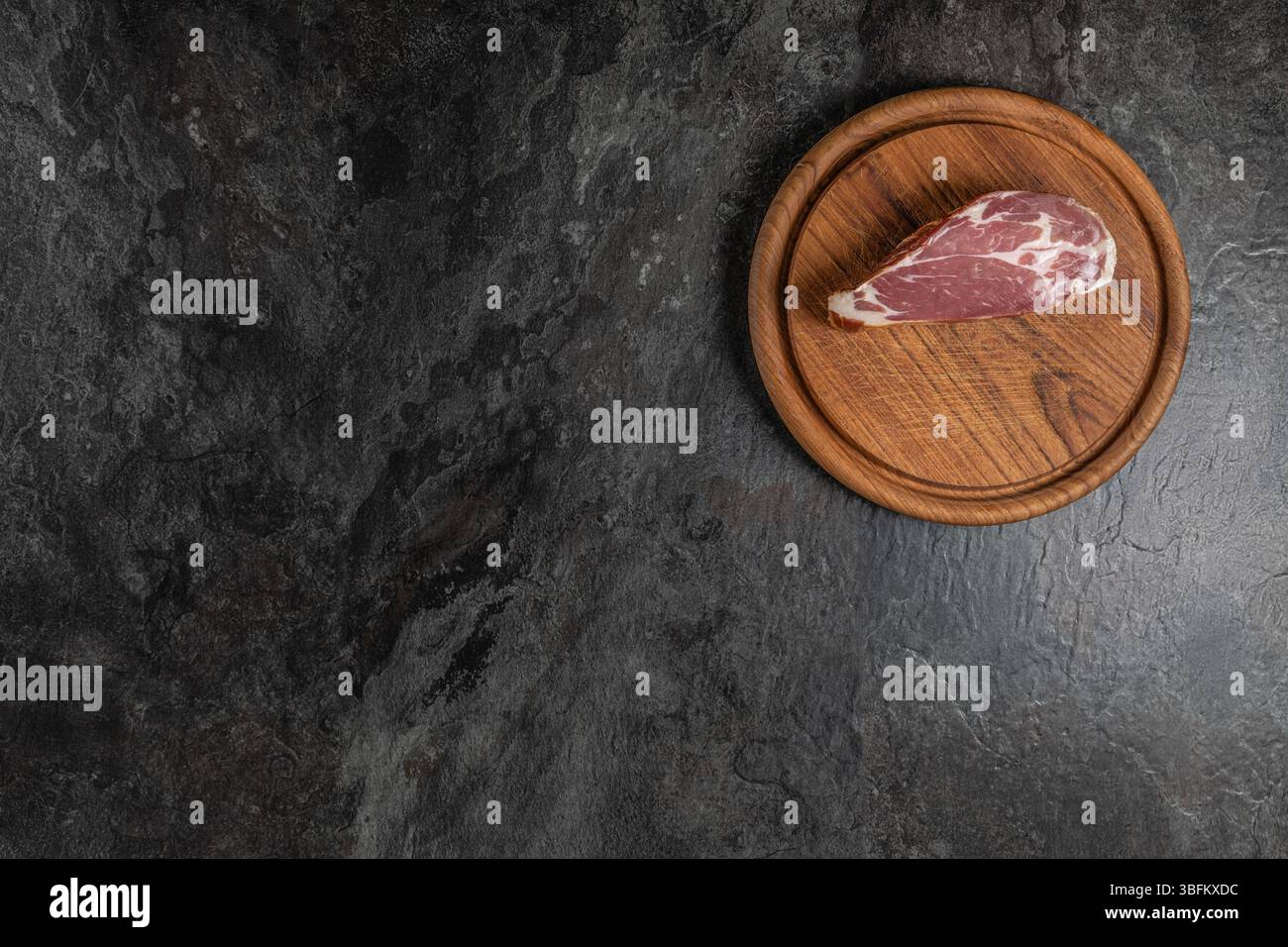 Prosciutto intero grezzo su tagliere in legno posizionato su una superficie di ardesia scura, illuminazione da studio con spazio per copiare a sinistra per testo o design Foto Stock