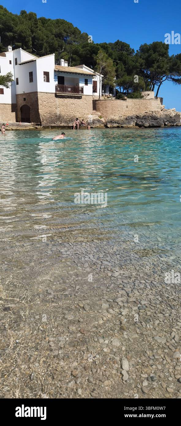 Cala Gat, Maiorca, Spagna; giugno 2025: Una tradizionale casa mediterranea si erge pacificamente tra le acque turchesi e la morbida sabbia rosa, incorniciata da pini Foto Stock