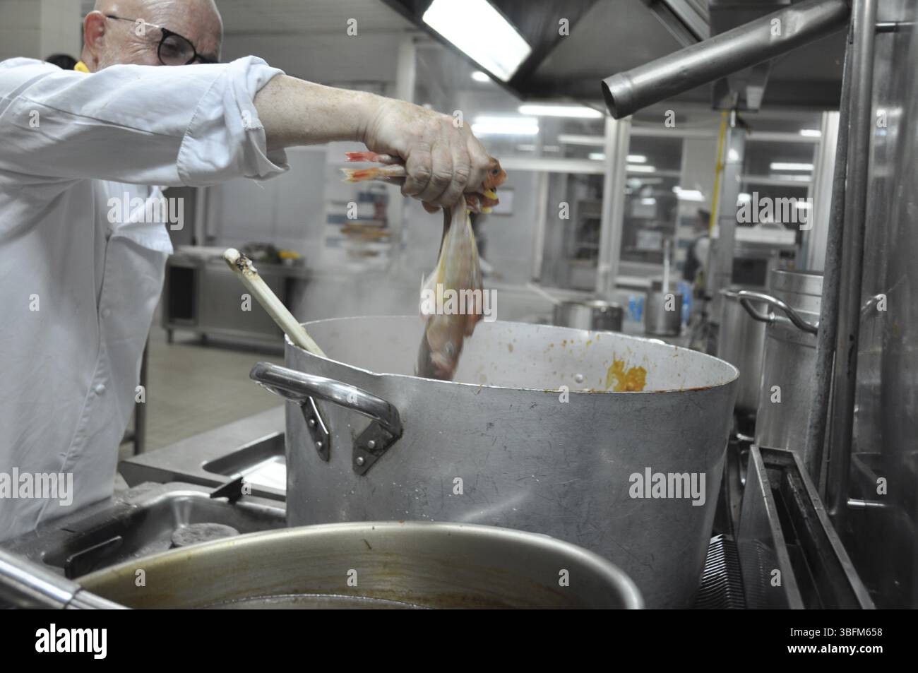 Chef che tiene il pesce sopra una pentola fumante in una cucina professionale, cuochi e chef preparano la cena con la tipica dieta Mediterranea in un resort di lusso Foto Stock